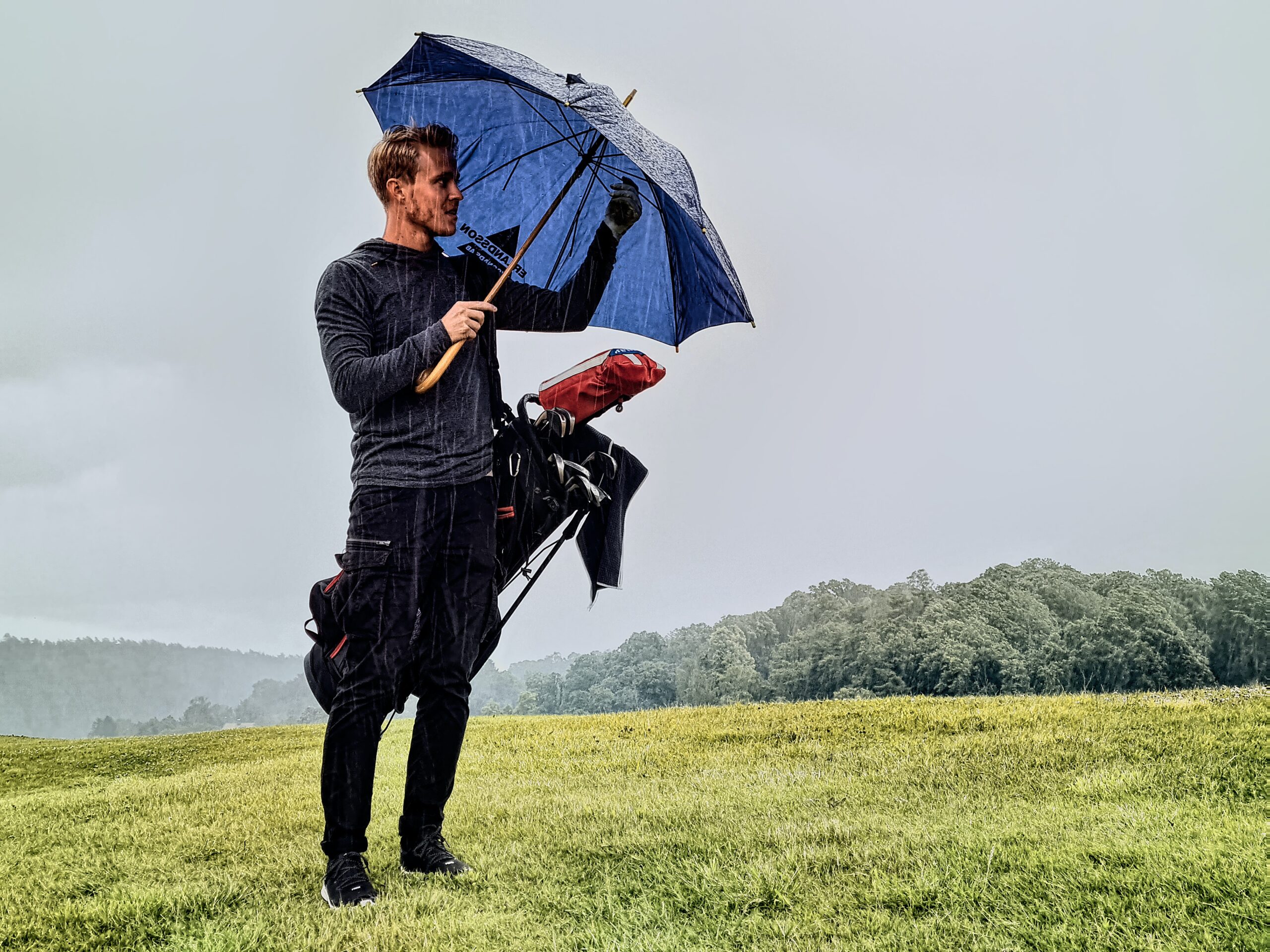 A golfer holding an umbrella and his golf bag in the rain.