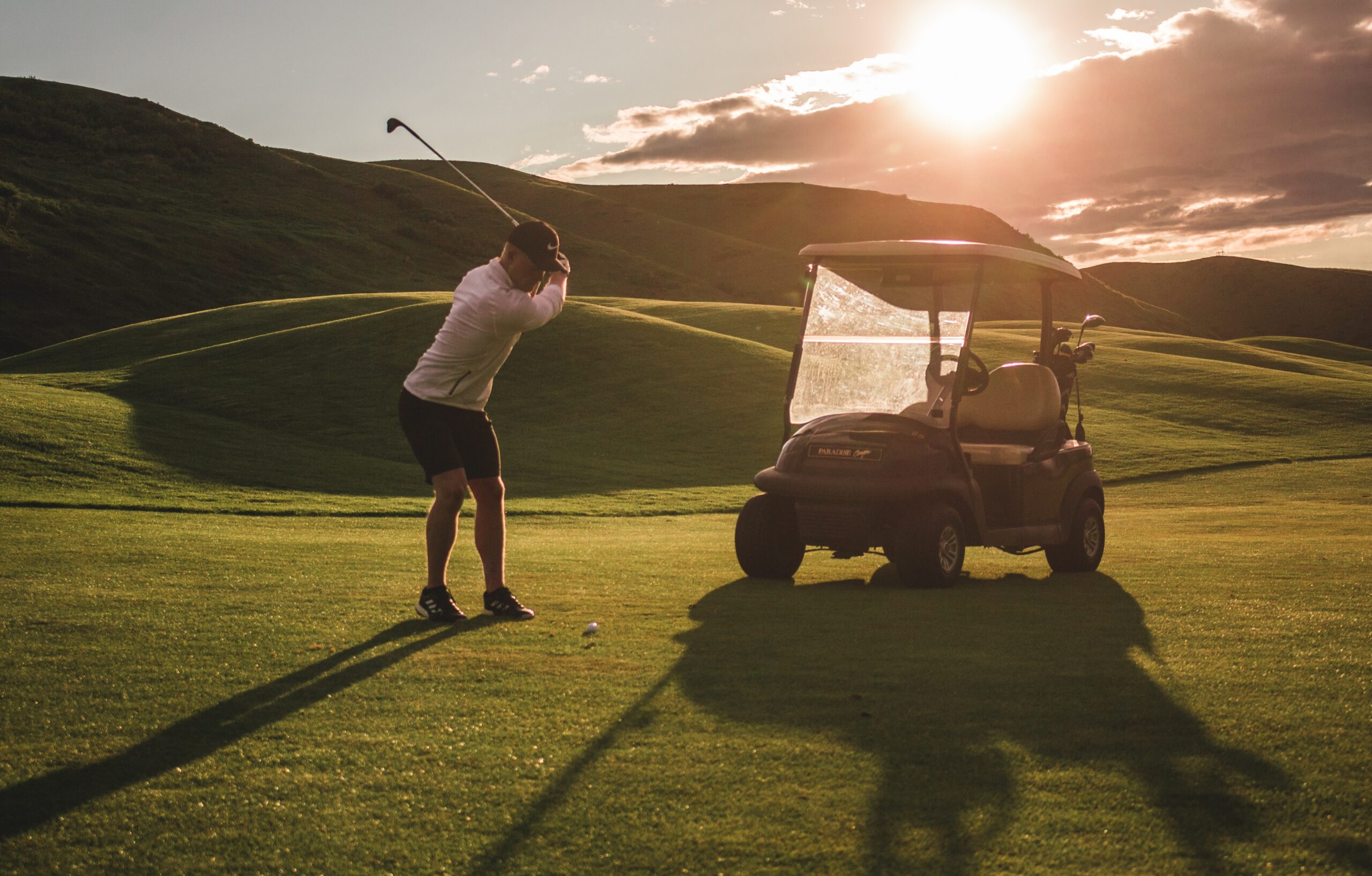 A golfer in mid-swing beside a golf cart as the sun sets in the background.