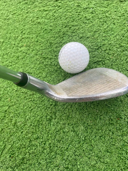 A high-lofted iron resting behind a golf ball on a green golf mat.