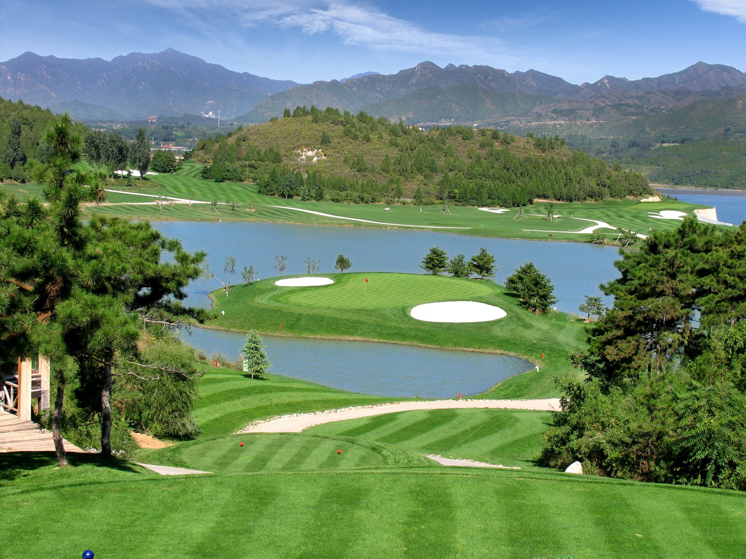 A golf course with a green jutting out into a river with mountains in the background.